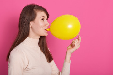 Side view of funny young woman with long dark hair inflating yellow balloon isolated over pink background, adorable female wearing rosy shirt, copy space for your advertisment or promotion text.