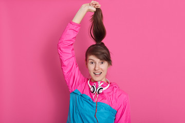 Studio shot of young Caucasian girl with dark hair holding her ponitail by hands playing with it, smiles, keeps mouth opened, lady with pleasant appearance posing isolated over pink background.
