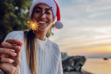 Attractive caucasian brunette in white sweater and with santa hat on head sitting on shore and...