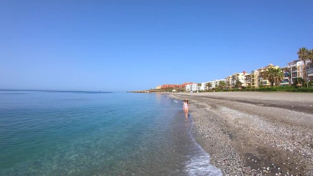 Aerial shot of a young woman in a swimsuit with a hat walking along the beach at the seashore