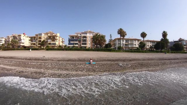 Aerial shot of a young woman in a swimsuit with a hat sitting on the shore of the beach.