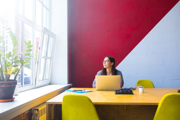Pensive female freelance social media content writer thinking and looking in window while sitting at workplace with pc laptop computer in modern co-working interior. Blogger dreaming