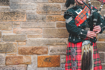 EDINBURGH, SCOTLAND, 24 March 2018 , Scottish bagpiper dressed in traditional red and black tartan dress stand before stone wall. Edinburgh, the most popular tourist city destination in Scotland.. - obrazy, fototapety, plakaty