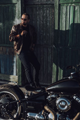 Fototapeta na wymiar Attractive biker with cigar standing near garage outdoors