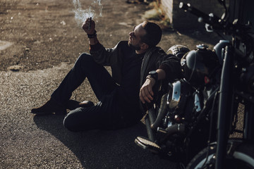 Naklejka na ściany i meble Handsome biker sitting near motorbike and smoking