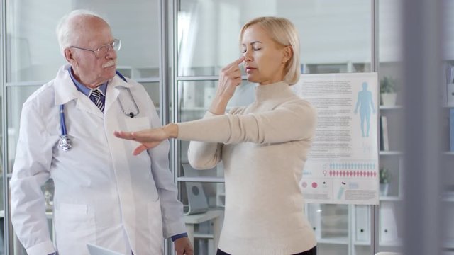 Senior neurologist talking to middle aged caucasian woman standing with eyes closed and then touching nose and clenching fists while having proprioception test during medical checkup