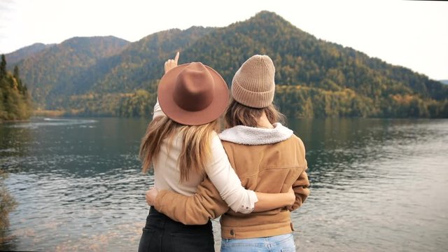 two attractive girls young women fashionably dressed tith hat on vacation on a lake in the mountains looking at the stunning scenery of nature. the camera floats forward . lesbian women