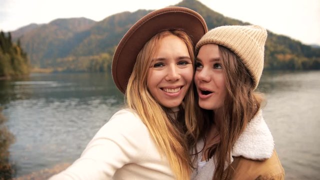 two very happy attractive girls young women fashionably dressed on vacation on a lake in the mountains shoot video chat, selfie and smile. the view from the camera. lesbian love. advertising, friends