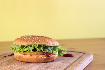 Homemade breaded chicken burger with lettuce on wooden chopping board with copy space on yellow background. Ketchup on cutting board.  
