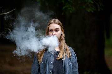 Vape teenager. Young white addict caucasian teenage girl with bruises under the eyes in casual clothes vaping electronic cigarette with cannabis liquid in the dark forest in the autumn evening.