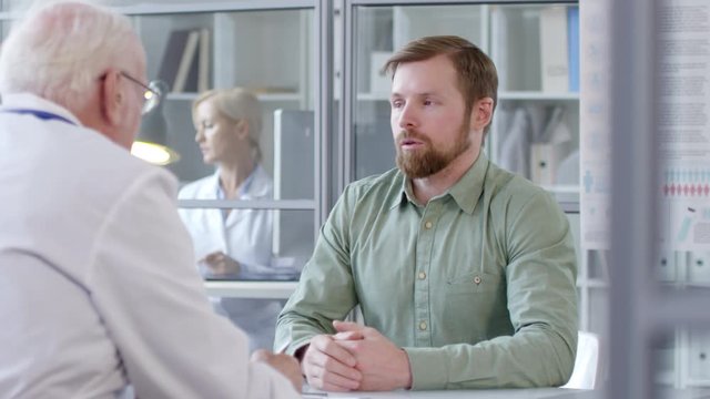 Young caucasian man complaining of sore throat and headache to senior physician while he taking notes during healthcare consultation in clinic