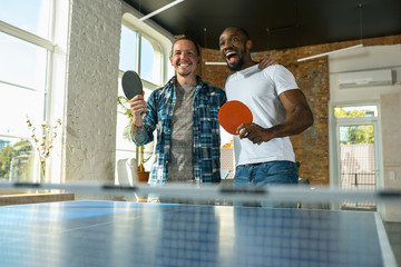Young men playing table tennis in workplace, having fun. Friends in casual clothes play ping pong...