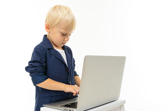 Little boy with short blonde hair, blue eyes, cute appearance, in white jacket, grey pants, blue jocket, standing with laptop and typing