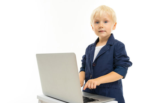 Little boy with short blonde hair, blue eyes, cute appearance, in white jacket, grey pants, blue jocket, standing with laptop and typing