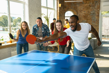 Young people playing table tennis in workplace, having fun. Friends in casual clothes play ping pong together at sunny day. Concept of leisure activity, sport, friendship, teambuilding, teamwork.