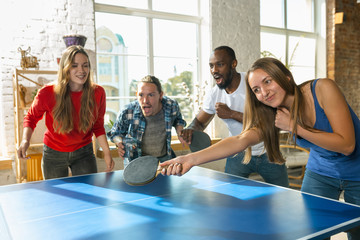 Young people playing table tennis in workplace, having fun. Friends in casual clothes play ping pong together at sunny day. Concept of leisure activity, sport, friendship, teambuilding, teamwork. - obrazy, fototapety, plakaty