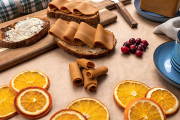 Fototapeta na wymiar Closeup shot of toasted breead with norwegian brown cheese/brunost and butter. Decorated with dried oranges and cranberries, wooden cutting board and blue coffee saucers. 