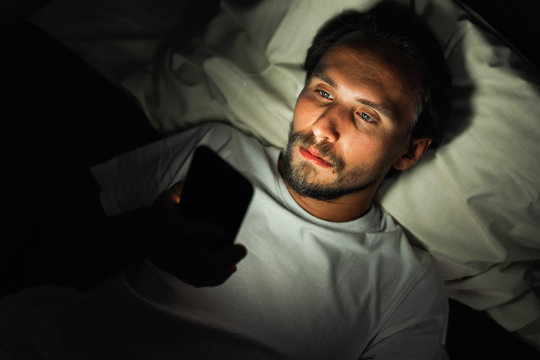 Young Handsome And Tired Man With A Beard Cannot Sleep And Is Watching Something On His Phone At Night.