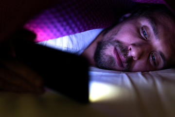 Bearded young man is lying in bed under his blanket. He cannot sleep and is watching something on...