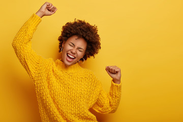 Curly haired girl in winter yellow sweater dances with arms spreading in air, enjoys music, has...