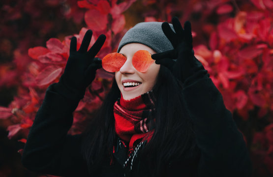 A young smiling girl in a gray hat, a dark coat and a plaid scarf covers her eyes with red and yellow leaves and smiles happily. The concept of a positive autumn mood.