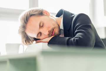 Handsome young man sleeping on pile of books at work desk. Coffee cup visible on a desk next to him.