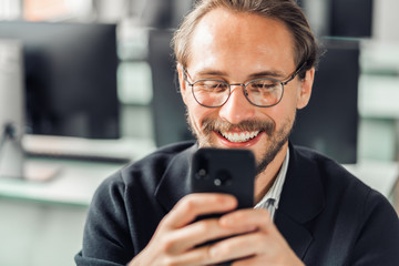 Young handsome man in glasses is interacting with his mobile phone while being at work. Happiness and content at work and being always reachable concepts.