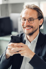 Young handsome man in glasses is interacting with his mobile phone while being at work. Happiness and content at work and being always reachable concepts.