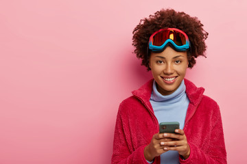 Indoor shot of pretty skier woman uses smartphone device for sending message, being on winter...