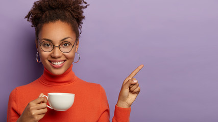 Adorable curly haired woman with tender smile, relaxes with drink in white mug, points on copy space, enjoys refreshing coffee, wears optical glasses and turtleneck, stands against purple background - Powered by Adobe