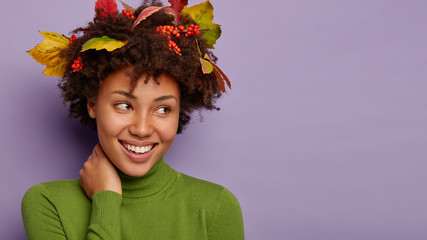 Natural beauty and happy autumn season concept. Close up shot of cheerful woman focused aside, celebrates fall, has colorful leaves in curly hair has charming smile isolated on purple wall blank space