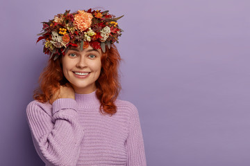 Adorable red haired woman has gentle smile, touches neck, healthy skin, wears handmade autummn wreath, knitted purple jumper, stands against violet wall with empty space. Season, beauty, emotions