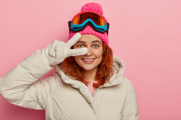 Smiling female tourist snowboards in mountains, wears winter hat and jacket, white gloves, makes...