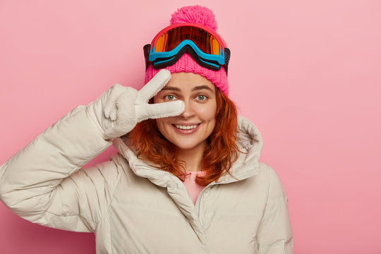 Smiling female tourist snowboards in mountains, wears winter hat and jacket, white gloves, makes peace gesture over eye, has seasonal rest, isolated on pink wall. Winter time and tourism concept