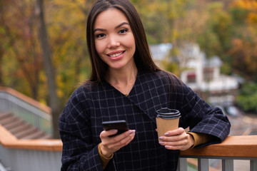 Young beautiful smiling woman in coat with coffee to go and cellphone happily looking in camera in park