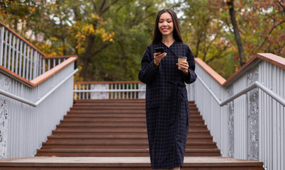 Young attractive casual woman in coat with coffee joyfully using cellphone while walking in city park