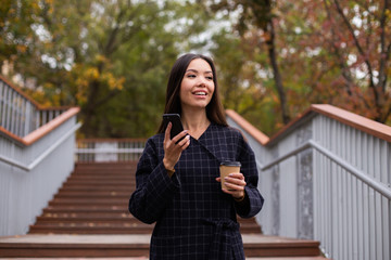 Young pretty casual woman in coat with coffee to go and cellphone happily looking away in park