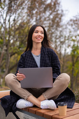Young beautiful casual woman happily working with laptop and coffee to go in city park