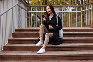 Young beautiful casual woman with coffee to go happily using cellphone while sitting on stairs in park