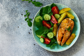 Vegetarian lunch or dinner, healthy eating concept. Grilled salmon, rustic baked potatoes and salad with fresh vegetables on a stone countertop. Top view flat lay background with copy space.
