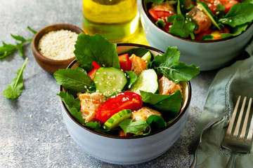 Tasty and healthy Salmon Grilled Salad with cucumber, tomatoes, fresh arugula and sesame seeds on a gray stone table.