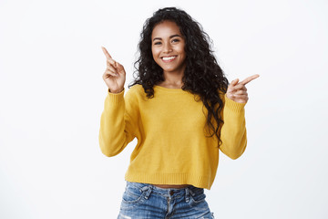 Cheerful friendly-looking attractive smiling african-american girl in yellow sweater pointing...