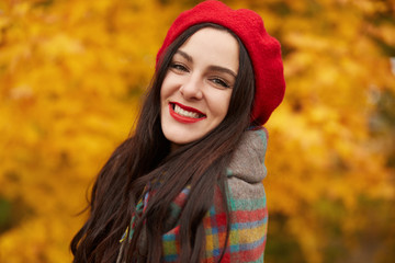 Happy brunette female in Autumn Park, young lady having fun outdoors surrounded with yellow trees and leaves, smiling woman posing outside, wearing red beret and wrapped warm checkered blanket.