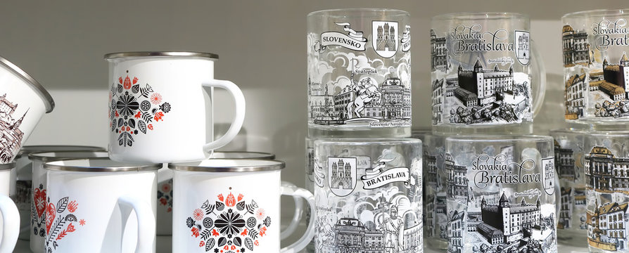 BRATISLAVA, SLOVAKIA - SEPTEMBER 01, 2019: White enamel mugs with pattern and glass cups with pictures of mail sightseeing places of Bratislava in souvenir shop. Banner