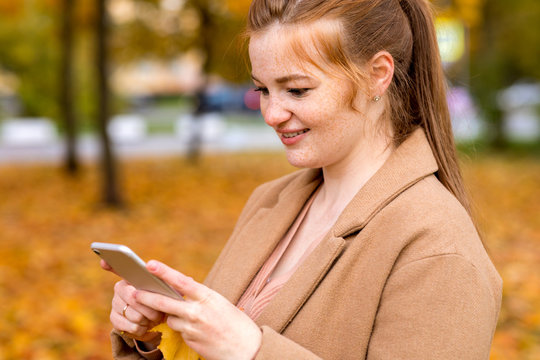 Image of young female using smartphone in the autumn park with colorful fallen leaves