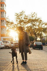 Portrait of a stylish full-length girl walking with a dog on a leash down the street on sunset...