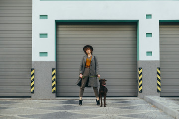 Fashionable girl in a hat and with curly hair holds a leash with a dog in her hand and poses at the camera against a gray wall background. Street portrait of stylish lady owner with dog on a walk.