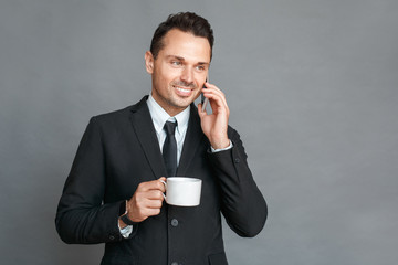 Businessman studio standing isolated on grey drinking coffee talking on smartphone smiling happy