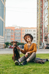 Beautiful girl in a hat and with a dog sitting on a green lawn, the girl holds a puppy on a leash and looks into the camera. Portrait of owner with cute brown dog on cityscape background.