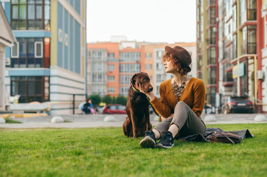 Portrait of a stylish girl in a hat sits with a beautiful brown dog on the lawn against the backdrop of a modern apartment building, looks at the puppy and strokes. Vacation with your pet.